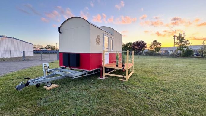 Tiny House "Red One", mit rotem Streifen und Runddach steht auf einem PKW Trailer.