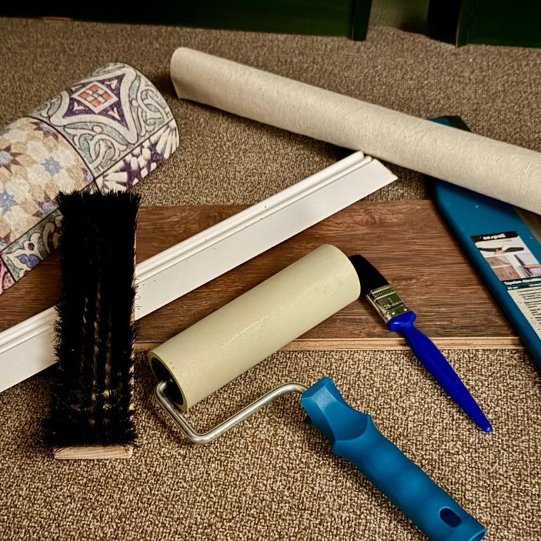 Tapetenrollen, Pinsel, Tapezierbürste und -rolle liegen mit einem Stück Laminat auf einem Teppich.