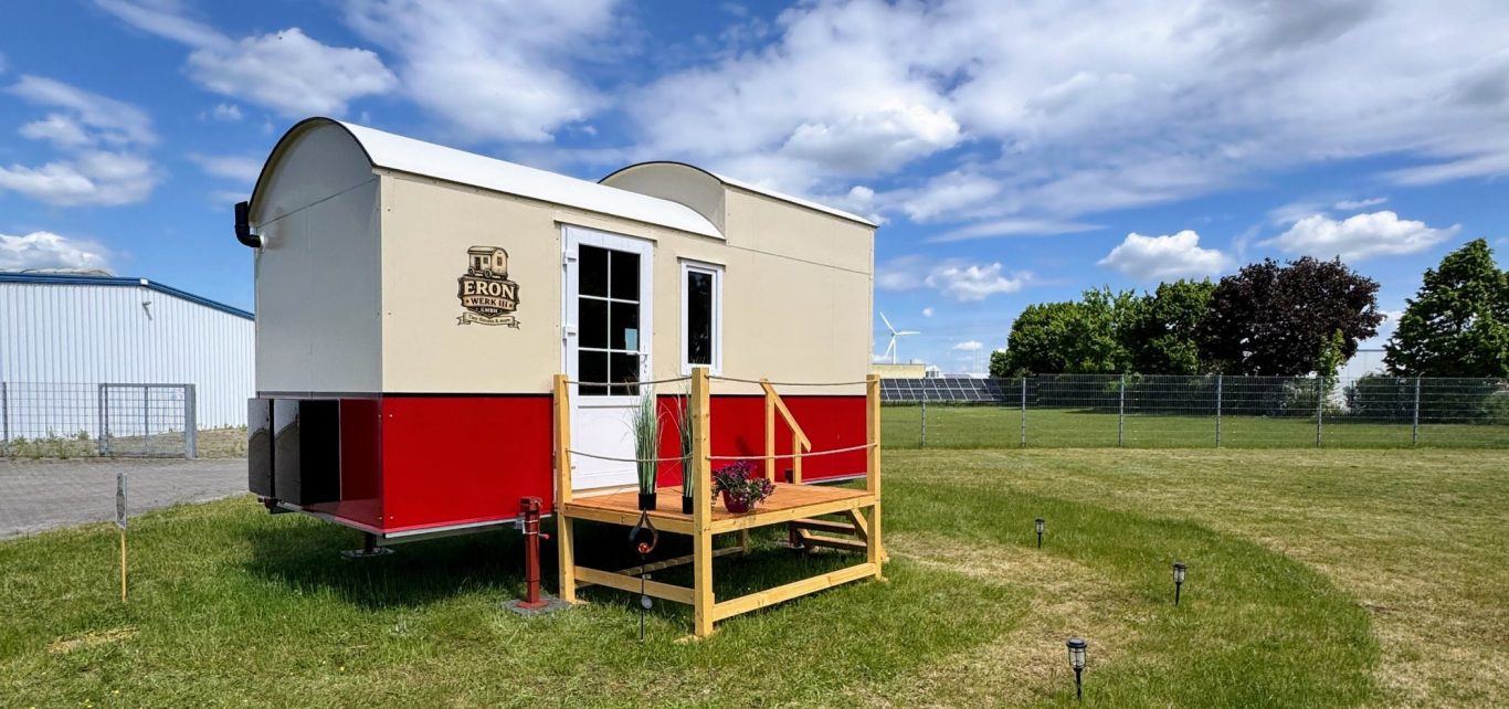 Mobiles Tiny House "Red One" mit Runddach, rotem Akzentstreifen und Holzterrasse davor.
