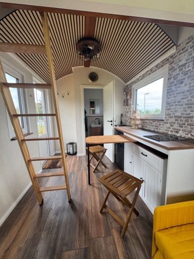 Ein Innenbild des Tiny Houses von der Wohnraumseite aus. Die Leiter, zwei Hocker und die Küchenzeile sind zu sehen.